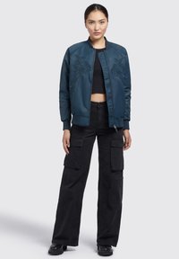 Teal bomber jacket dengan desain bordir bunga, ritsleting depan, manset bergaris, dan panjang crop, dipasangkan dengan celana cargo hitam.