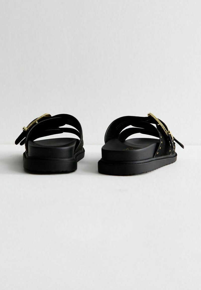 Sandalias negras con correas ajustables, hebillas en tono dorado, un acabado texturizado y suelas planas. Presenta un diseño minimalista y sin adornos.