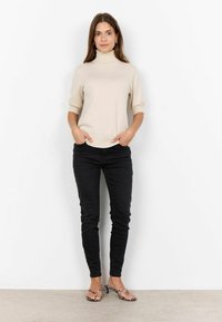 Beige garbó pulóver rövid ujjakkal, fekete szűk farmernadrággal és mintás papuccsal, fehér háttér előtt áll.