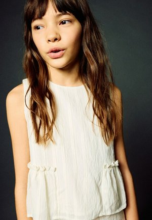 Jeune fille aux longs cheveux bruns portant une robe sans manches blanche texturée, regardant légèrement sur le côté sur un fond sombre.