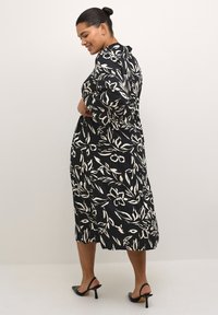 Schwarzes knielanges Kleid mit weißem Blumenmuster, aus glattem Stoff; verfügt über einen Kragen und Dreiviertelärmel, kombiniert mit schwarzen Sandalen mit Absatz.