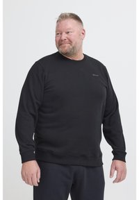Uomo con capelli biondi corti e barba, vestito con una felpa nera e pantaloni, che guarda a sinistra davanti a uno sfondo neutro.