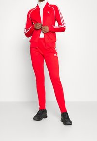 Röd träningsoveralljacka och byxor med vita ränder, med dragkedja och logotypdetalj. Kombinerad med svarta sportskor.