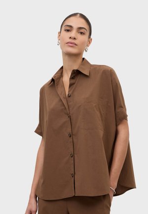 Femme aux cheveux tirés en arrière, portant des boucles d'oreilles argentées et une chemise marron à manches courtes et boutons, ample, avec une poche avant, la main dans la poche.