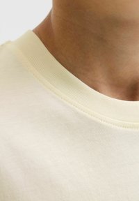 Pogled izbliza na vrat osobe i ovratnik majice s okruglim izrezom u krem boji, s finom rebrastom teksturom i glatkom tkaninom.