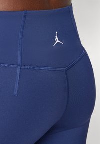Tamsiai mėlyni sportiniai leggingai su lygiu paviršiumi, su baltu Jumpman logotipu ant nugaros ir kontrastingais siuvinėjimo detalėmis.