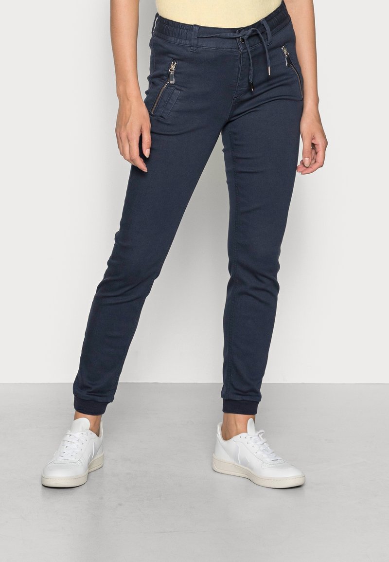 Femme portant un pantalon de jogging bleu marine avec des poches zippées et des baskets blanches, se tenant devant un fond clair.