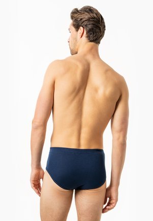 Mann mit kurzen dunklen Haaren, von hinten gesehen, trägt dunkelblaue Slips vor einem schlichten weißen Hintergrund.