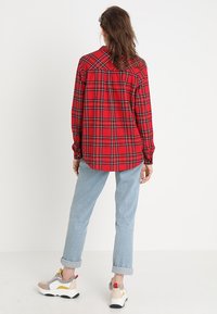 Person med langt hår iført en rød plaidskjorte, lyseblå opbukede jeans og multikolore sneakers stående foran en ensfarvet hvid baggrund.
