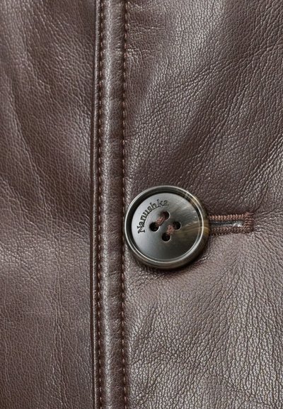 Îmbrăcăminte din piele maro, prezentând un prim-plan al unui buton rotund gravat, cu patru găuri și detalii textuale, completat de cusături vizibile.