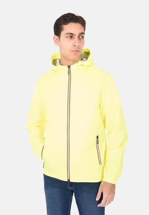 Giovane uomo con giacca con cappuccio gialla brillante con cerniera frontale e jeans blu, in piedi con le mani nelle tasche, che guarda di lato.