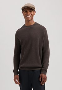 Brun ribbet genser med rund hals og lange ermer, brukt med mørke bukser. En lys grå flat cap er brukt på hodet.