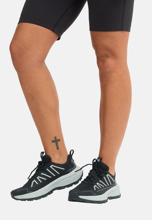 Benën met zwart-witte sportsneakers en zwarte shorts, met een kleine kruis-tatoeage op de rechterenkel tegen een witte achtergrond.