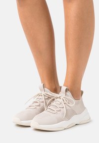 Zapatillas deportivas de color beige claro, fabricadas con material de malla y punto. Cuentan con una suela de goma, diseño de cordones y un cuello acolchado.