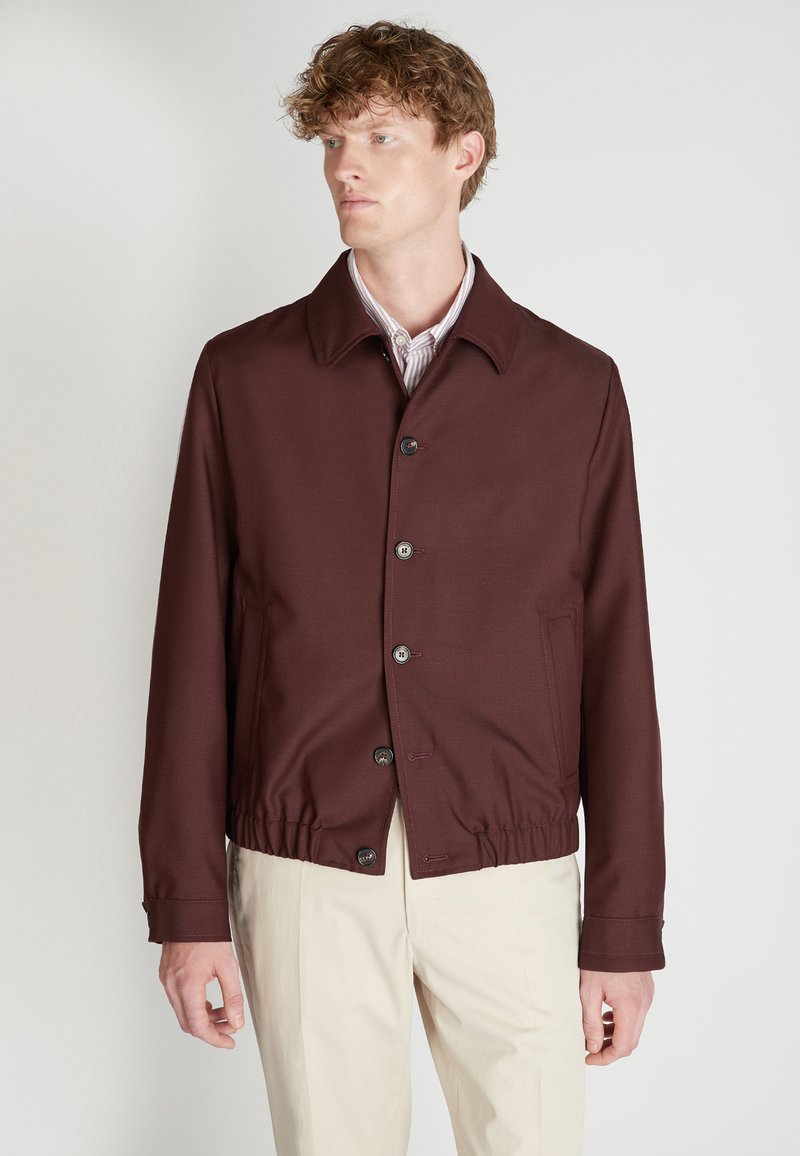 Chaqueta burdeos con cuello tipo camisa, cierre frontal con botones y cintura elástica. Presenta bolsillos laterales y una textura de tela suave.