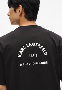 Melnas kokvilnas T-krekls ar baltu drukātu tekstu mugurā: "KARL LAGERFELD PARIS 21 RUE ST-GUILLAUME." Standarta apkakles dizains.