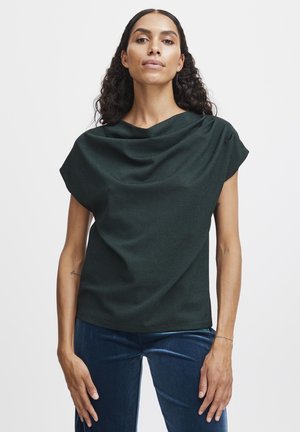 Blusa verde scuro con scollatura drappeggiata, maniche corte e un tessuto morbido e texturizzato. Abbinata a pantaloni in velluto blu a vita alta.