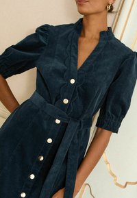 Vestito in corduroy blu scuro con maniche a sbuffo corte, scollatura a scallop e chiusura frontale con bottoni dorati. Include una cintura coordinata in tessuto.