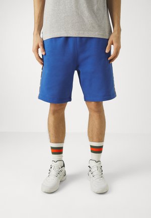 Pantalones cortos deportivos azules de corte relajado, con franjas laterales y una textura suave. Combinados con zapatillas blancas y calcetines multicolores.
