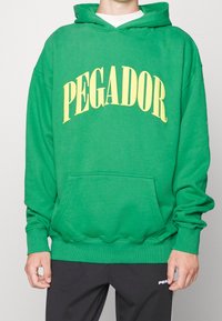 Una persona indossa una felpa verde brillante con la scritta "PEGADOR" in bold giallo, abbinata a pantaloni scuri. Lo sfondo è neutro.