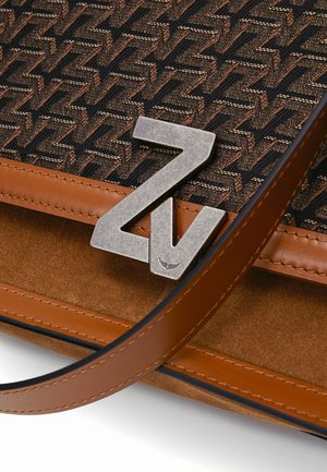 Personne portant un haut noir sans manches et un pantalon en cuir marron, tenant un sac à main marron à motifs avec un fermoir argenté "Zv", assise sur une chaise blanche.