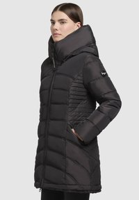 Doudoune noire avec un col haut et ajustable, motif matelassé, deux poches latérales et une finition lisse et brillante. Fermeture frontale zippée.