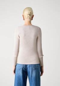 Personne aux cheveux blonds attachés en chignon, portant des boucles d'oreilles argentées en forme d'anneau, un haut côtelé beige à manches longues et un jean bleu, se tenant devant un fond blanc.