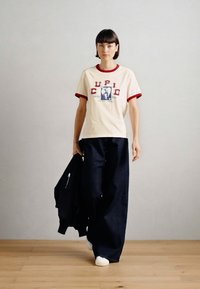 Κρέμα μπλουζάκι με κόκκινο περιθώριο και γραφικά σχέδια, συνδυασμένο με φαρδιά τζιν παντελόνια σε σκούρο χρώμα και λευκά αθλητικά παπούτσια. Το μοντέλο κρατά μια μαύρη ζακέτα.