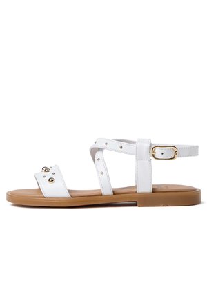 Sandale plate blanche avec semelle beige, munie d’une bride à la cheville et de clous dorés sur plusieurs brides ainsi qu’une fermeture à boucle dorée.