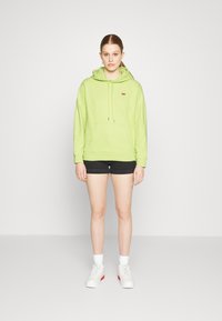 Lys grønn hettegenser med frontlomme og snøring, kombinert med svarte shorts og hvite joggesko med røde detaljer.