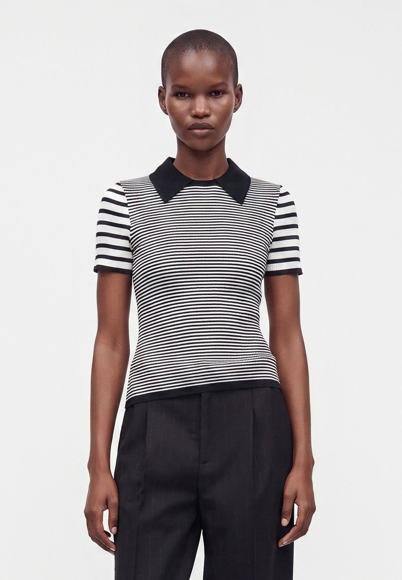 Femme portant un haut à manches courtes rayé noir et blanc avec un col noir et un pantalon noir, debout devant un fond blanc uni.