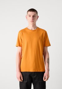 Orange bomulds T-shirt med korte ærmer, rund halsudskæring og en lille logo detalje på brystet; iført med mørke denim bukser.