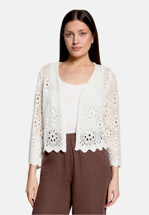 Kvinde med glat mørkt hår iført en hvid hæklede cardigan over en hvid top og løse brune bukser, stående mod en ensfarvet baggrund.