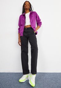Jachetă violeta cu fermoar, tricou alb scurt, blugi negri cu picior drept și adidași albi cu accente verde neon. Material neted și croială casual.