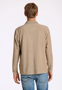 Camicetta beige a maniche lunghe con colletto, caratterizzata da una vestibilità rilassata e tessuto testurizzato, abbinata a jeans blu. Dettagli delle cuciture visibili.