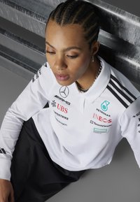 Vit polo t-shirt med svarta accenter och sponsorlogotyper. Har krage, långa ärmar och ett texturerat tyg. Svarta byxor under.