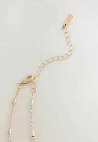 Goldene Kettenaccessoires mit verstellbaren Gliedern, einem Karabinerhaken und zylindrischen Akzenten. Glatte Textur und polierte Oberfläche.