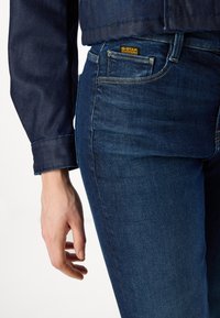 Jeans en denim bleu foncé avec une coupe slim. Les caractéristiques incluent un logo G-Star jaune, des coutures contrastées et une texture subtile. Main positionnée à côté des jeans.