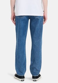 Vaqueros de mezclilla azul con un corte relajado, que cuentan con un ligero desgaste y cinco bolsillos. Combinados con zapatillas negras y una camisa blanca.