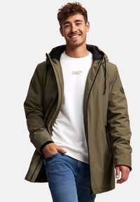 Veste imperméable vert olive avec capuche, doublure noire, poches zippées et cordons de serrage réglables. Portée par-dessus un t-shirt blanc et un jeans.