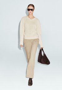 Beige gebreide trui met een V-hals, tan kleurige broek en een donkerbruine suède tas. Het model draagt oversized zwarte zonnebrillen en een paar bruine instappers.