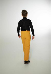 Žluté kalhoty se zadní kapsou, černé tričko s dlouhým rukávem, černé šle a černé boty. Jednoduchý, rovný střih, minimalistický styl.