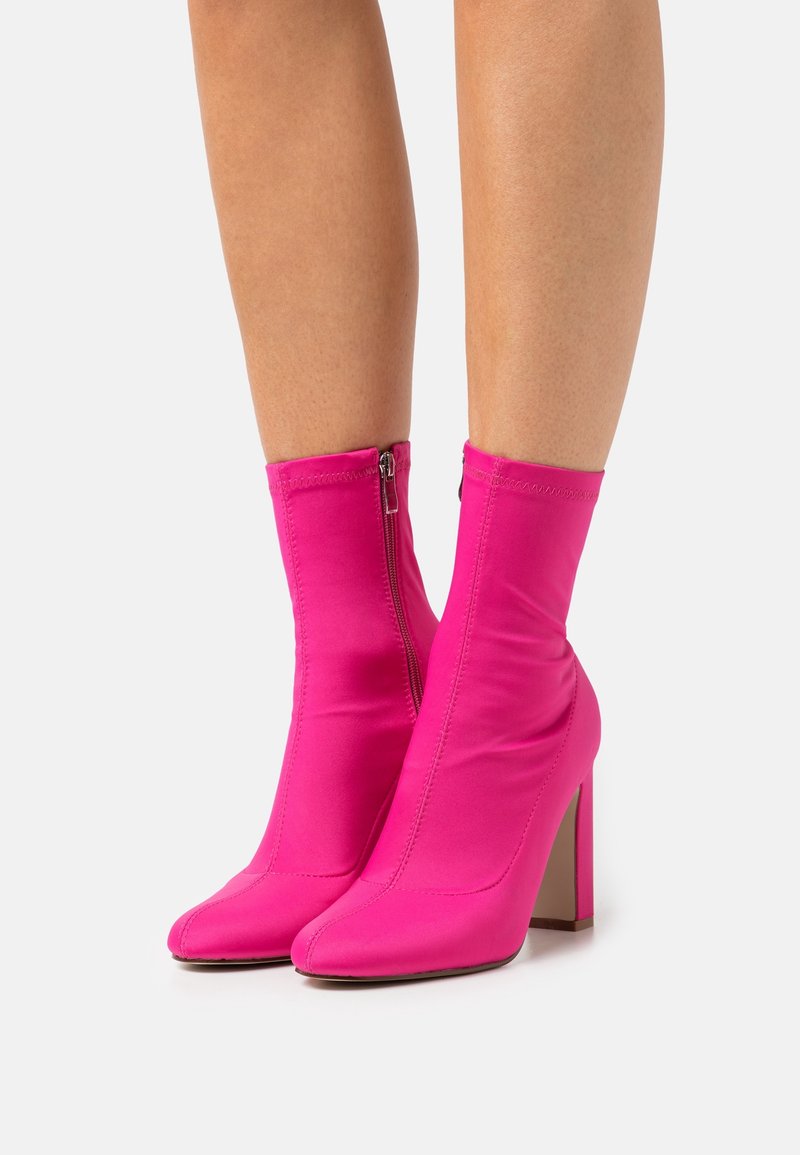 Jambes portant des bottines rose vif à bouts pointus, talons carrés et fermetures éclair latérales sur un fond blanc uni.