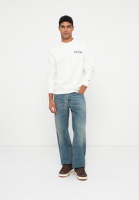 Muž stojící s rukou v kapse, oblečený v bílém svetru s červenobílým logem, světlých modrých širokých džínách a hnědých teniskách na bílém pozadí.