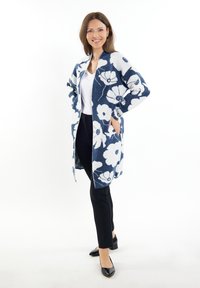 Cardigan lungo a fantasia floreale in blu navy e bianco, tessuto lavorato a maglia, apertura frontale, tasche, abbinato a una maglietta bianca e pantaloni neri.