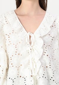 White embroidered blouse with floral patterns, ruffled neckline, and tie closure. Features eyelet detail and flowing sleeves.