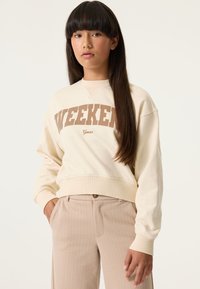 Sudadera color crema con la palabra "WEEKEND" en letras marrones, con cuello redondo y puños acanalados. Combinada con pantalones beige a rayas.