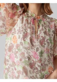 Blouse en chiffon à motif floral avec un col rond, fermeture par un nœud et manches courtes bouffantes, dans des couleurs pastel rose, vert et orange.