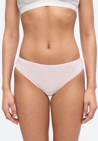 Žena oblečená v svetloružovom bavlnenom spodnom prádle a svetlosivej športovej podprsenke, stojaca pred jednoduchým bielym pozadím.