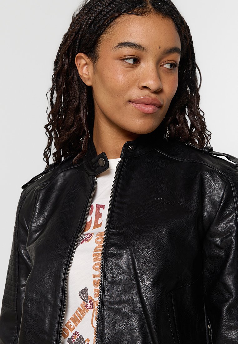 Joven mujer con cabello rizado que lleva una chaqueta de cuero negra y una camiseta gráfica blanca, mirando ligeramente hacia un lado.